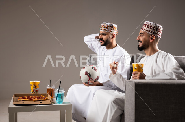 تناول الوجبات السريعة والمشاريب المنعشة, التفاعل والاندماج بمشاهدة الفريق المفضل, المباريات والالعاب الرياضية, ايماءات الفرح والحماس بالفوز والانتصار, بورتريه لرجلان عربيان عمانين خليجيان يرتديان الدشداشة والكمة احدهما يمسك بيده كرة قدم, الشغف بالانشطة الترفيهية, خلفية رمادية