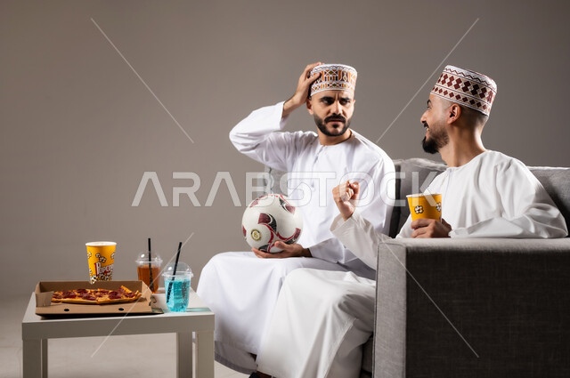 تناول الوجبات السريعة والمشاريب المنعشة, التفاعل والاندماج بمشاهدة الفريق المفضل, المباريات والالعاب الرياضية, ايماءات الفرح والحماس بالفوز والانتصار, بورتريه لرجلان عربيان عمانين خليجيان يرتديان الدشداشة والكمة احدهما يمسك بيده كرة قدم, الشغف بالانشطة الترفيهية, خلفية رمادية