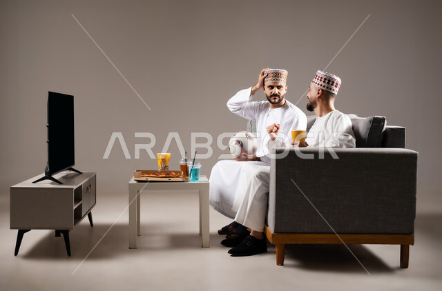 Interaction and integration by watching the favorite team, sports matches and games, gestures of joy and enthusiasm for victory and triumph, portrait of two Omani Gulf Arab men wearing a dishdasha and a kabbah, one of them holding a football in his hand, passion for recreational activities, gray background