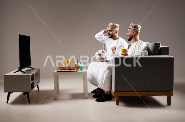 Interaction and integration by watching the favorite team, sports matches and games, gestures of joy and enthusiasm for victory and triumph, portrait of two Omani Gulf Arab men wearing a dishdasha and a kabbah, one of them holding a football in his hand, passion for recreational activities, gray background