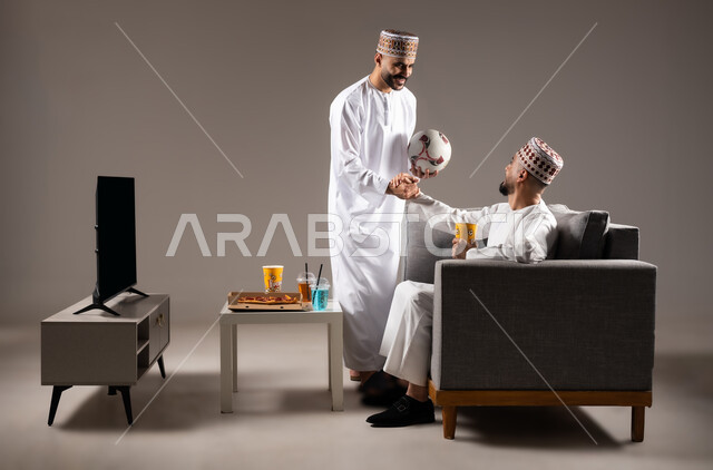 Interaction and integration by watching the favorite team, sports matches and games, gestures of joy and enthusiasm for victory and triumph, portrait of two Omani Gulf Arab men wearing a dishdasha and a kabbah, one of them holding a football in his hand, passion for recreational activities, gray background