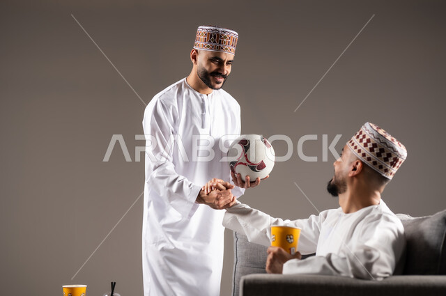 تناول الوجبات السريعة والمشاريب المنعشة, التفاعل والاندماج بمشاهدة الفريق المفضل, المباريات والالعاب الرياضية, ايماءات الفرح والحماس بالفوز والانتصار, بورتريه لرجلان عربيان عمانين خليجيان يرتديان الدشداشة والكمة احدهما يمسك بيده كرة قدم, الشغف بالانشطة الترفيهية, خلفية رمادية
