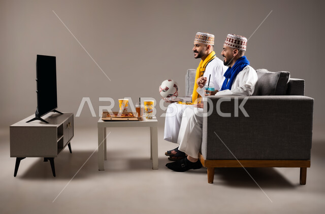 Interaction and integration by watching the favorite team, sports matches and games, gestures of joy and enthusiasm for victory and triumph, portrait of two Omani Gulf Arab men wearing a dishdasha and a kabbah, one of them holding a football in his hand, passion for recreational activities, gray background