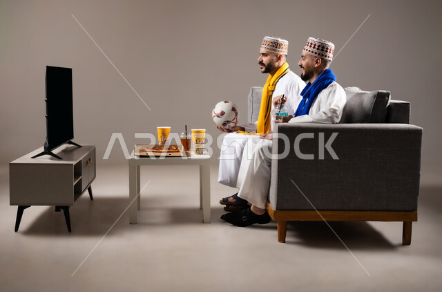 Interaction and integration by watching the favorite team, sports matches and games, gestures of joy and enthusiasm for victory and triumph, portrait of two Omani Gulf Arab men wearing a dishdasha and a kabbah, one of them holding a football in his hand, passion for recreational activities, gray background
