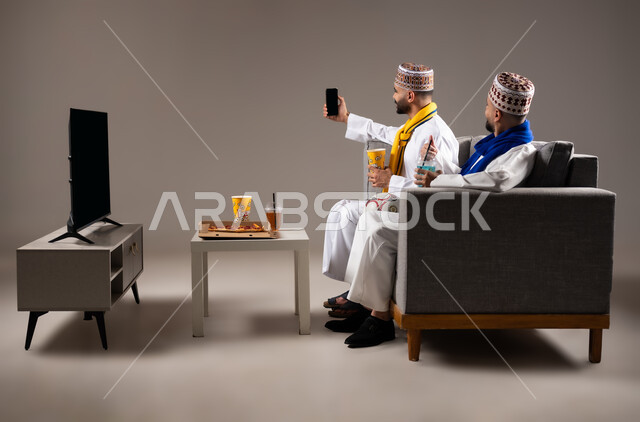 التقاط الصور التذكارية, تناول الوجبات السريعة والمشاريب المنعشة, قضاء وقت ممتع مع العائلة والاصدقاء, ايماءات الفرح والسرور والسعادة, بورتريه لرجلان عربيان عمانين خليجيان يرتديان الدشداشة والكمة يمسك احدهم هاتف محمول ويلتقط صورة, خلفية رمادية