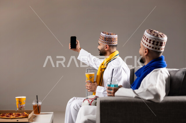 التقاط الصور التذكارية, تناول الوجبات السريعة والمشاريب المنعشة, قضاء وقت ممتع مع العائلة والاصدقاء, ايماءات الفرح والسرور والسعادة, بورتريه لرجلان عربيان عمانين خليجيان يرتديان الدشداشة والكمة يمسك احدهم هاتف محمول ويلتقط صورة, خلفية رمادية