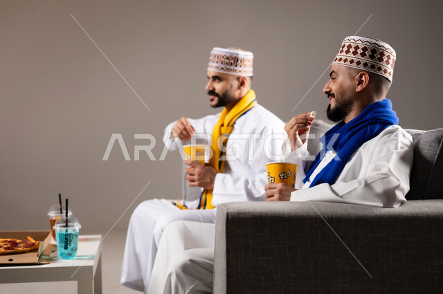 مفهوم التسليه والترفيه الجماعي, قضاء وقت ممتع بتناول الفوشار اثناء مشاهده التلفاز، بورتريه لصديقان عربيان خليجيان عمانيان يرتديان الكمه والدشداشة يجلسان على الأريكه يحملان بيدهما أكواب مليئه بالبوب كورن، تعابير وايماءات السعاده والسرور، خلفية رمادية