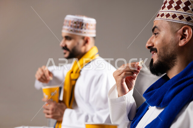 مفهوم التسليه والترفيه الجماعي, قضاء وقت ممتع بتناول الفوشار اثناء مشاهده التلفاز، بورتريه لصديقان عربيان خليجيان عمانيان يرتديان الكمه والدشداشة يجلسان على الأريكه يحملان بيدهما أكواب مليئه بالبوب كورن، تعابير وايماءات السعاده والسرور، خلفية رمادية