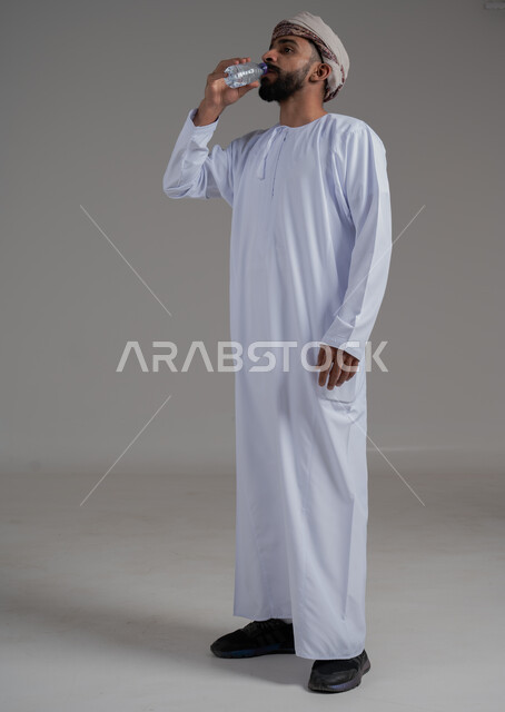أهمية شرب الماء النقي للمحافظة على بشرة نضرة، المحافظة على سلامة الجهاز المناعي، التخلص من سموم الجسم، الحفاظ على صحة الكلى ومعالجتها، بورتريه لرجل عربي خليجي عماني يرتدي الدشداشه والعمامة يقوم بشرب الماء م قارورة بلاستيكية، الجسم بالطول الكامل، خلفية بيضاء