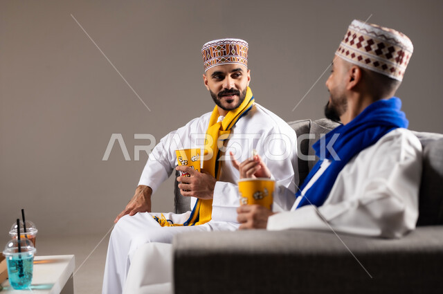 مفهوم التسليه والترفيه الجماعي, قضاء وقت ممتع بتناول الفوشار اثناء مشاهده التلفاز، بورتريه لصديقان عربيان خليجيان عمانيان يرتديان الكمه والدشداشة يجلسان على الأريكه يحملان بيدهما أكواب مليئه بالبوب كورن، تعابير وايماءات السعاده والسرور، خلفية رمادية