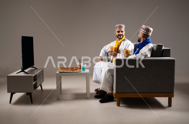 مفهوم التسليه والترفيه الجماعي, قضاء وقت ممتع بتناول الفوشار اثناء مشاهده التلفاز، بورتريه لصديقان عربيان خليجيان عمانيان يرتديان الكمه والدشداشة يجلسان على الأريكه يحملان بيدهما أكواب مليئه بالبوب كورن، تعابير وايماءات السعاده والسرور، خلفية رمادية