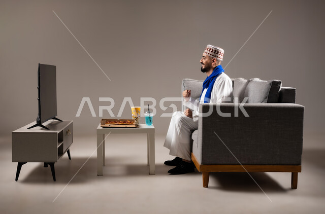 شرب العصائر والمشاريب الباردة والمنعشة اثناء مشاهده التلفاز، قضاء وقت ممتع, بورتريه لرجل عربي خليجي عماني يرتدي الدشداشة والكمه يجلس في غرفة المعيشة على الأريكه ويتناول البيتزا والفوشار، مفهوم التسليه والترفيه, خلفية رمادية