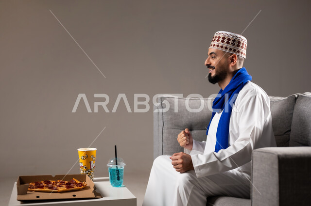 شرب العصائر والمشاريب الباردة والمنعشة اثناء مشاهده التلفاز، قضاء وقت ممتع, بورتريه لرجل عربي خليجي عماني يرتدي الدشداشة والكمه يجلس في غرفة المعيشة على الأريكه ويتناول البيتزا والفوشار، مفهوم التسليه والترفيه, خلفية رمادية