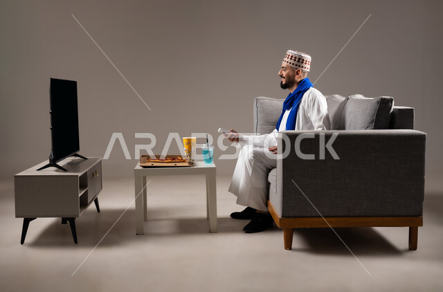 شرب العصائر والمشاريب الباردة والمنعشة اثناء مشاهده التلفاز، قضاء وقت ممتع, بورتريه لرجل عربي خليجي عماني يرتدي الدشداشة والكمه يجلس في غرفة المعيشة على الأريكه ويتناول البيتزا والفوشار، مفهوم التسليه والترفيه, خلفية رمادية