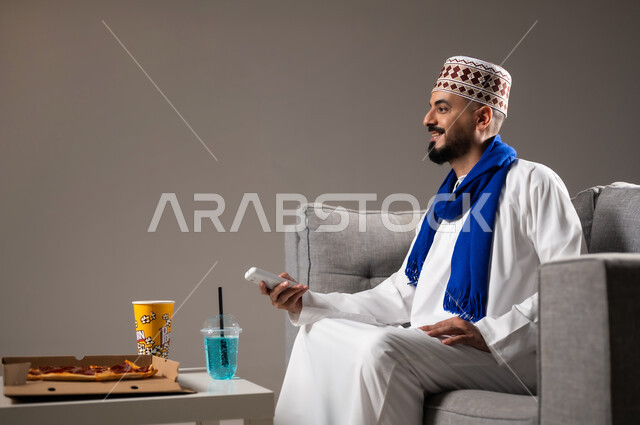 شرب العصائر والمشاريب الباردة والمنعشة اثناء مشاهده التلفاز، قضاء وقت ممتع, بورتريه لرجل عربي خليجي عماني يرتدي الدشداشة والكمه يجلس في غرفة المعيشة على الأريكه ويتناول البيتزا والفوشار، مفهوم التسليه والترفيه, خلفية رمادية