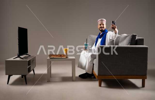 التقاط الصور التذكارية, شرب العصائر والمشاريب الباردة والمنعشة، قضاء وقت ممتع, بورتريه لرجل عربي خليجي عماني يرتدي الدشداشة والكمه يجلس في غرفة المعيشة على الأريكه يمسك بيده الهاتف المحمول ويقوم بتصوير نفسه، مفهوم التسليه والترفيه, خلفية رمادية