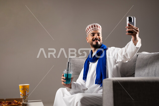 التقاط الصور التذكارية, شرب العصائر والمشاريب الباردة والمنعشة، قضاء وقت ممتع, بورتريه لرجل عربي خليجي عماني يرتدي الدشداشة والكمه يجلس في غرفة المعيشة على الأريكه يمسك بيده الهاتف المحمول ويقوم بتصوير نفسه، مفهوم التسليه والترفيه, خلفية رمادية