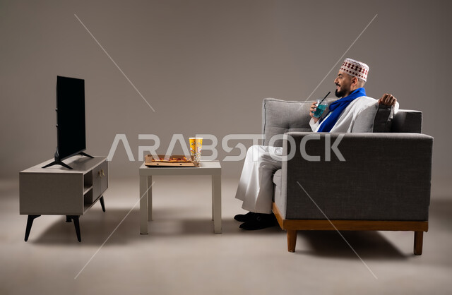 شرب العصائر والمشاريب الباردة والمنعشة اثناء مشاهده التلفاز، قضاء وقت ممتع, بورتريه لرجل عربي خليجي عماني يرتدي الدشداشة والكمه يجلس في غرفة المعيشة على الأريكه ويتناول عصير طبيعي، مفهوم التسليه والترفيه, خلفية رمادية