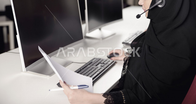 موظفة عربية خليجية سعودية تعمل في خدمة العملاء وتمسك الاوراق بيدها، موظفة ترتدي سماعة الاتصال، استعلامات واستفسارات، الاجابه على اسئلة العملاء، اجراء اتصال فيديو من خلال الحاسوب، متابعة وتطوير العمل
