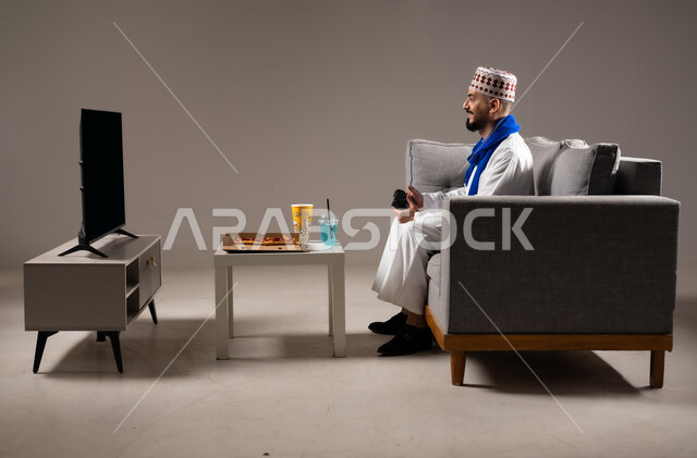 تناول وجبات خفيفة اثناء اللعب, قضاء وقت ممتع, بورتريه لرجل عربي خليجي عماني يرتدي الدشداشة والكمه يجلس في غرفة المعيشة على الأريكه ويمسك بيده عصا التحكم, لعب العاب الفيديو بإيماءات تدل على السعادة والسرور، المتعة و التسلية والتفاعل بالبلايستيشن الترفيهية, خلفية رمادية