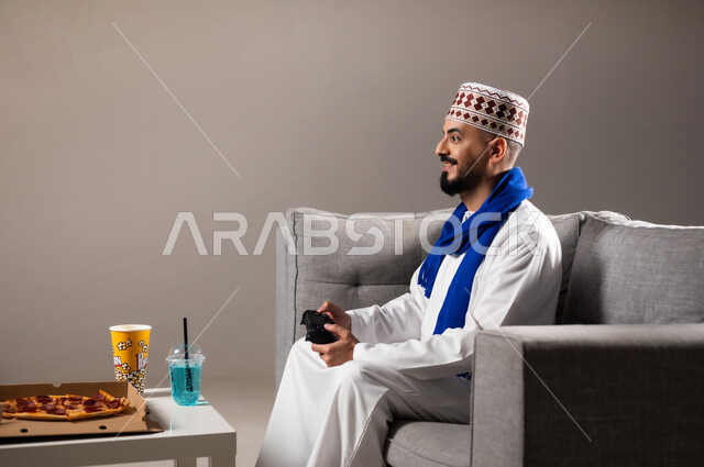 تناول وجبات خفيفة اثناء اللعب, قضاء وقت ممتع, بورتريه لرجل عربي خليجي عماني يرتدي الدشداشة والكمه يجلس في غرفة المعيشة على الأريكه ويمسك بيده عصا التحكم, لعب العاب الفيديو بإيماءات تدل على السعادة والسرور، المتعة و التسلية والتفاعل بالبلايستيشن الترفيهية, خلفية رمادية