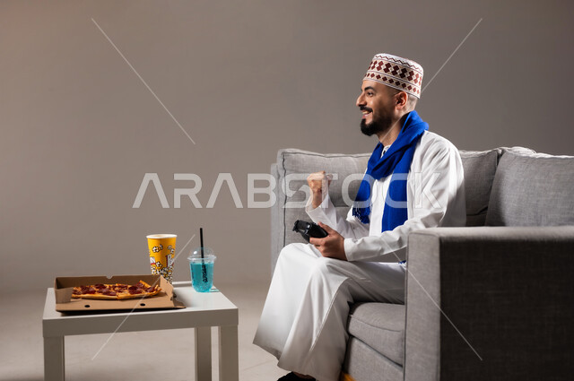 تناول وجبات خفيفة اثناء اللعب, قضاء وقت ممتع, بورتريه لرجل عربي خليجي عماني يرتدي الدشداشة والكمه يجلس في غرفة المعيشة على الأريكه ويمسك بيده عصا التحكم, لعب العاب الفيديو بإيماءات تدل على السعادة والسرور، المتعة و التسلية والتفاعل بالبلايستيشن الترفيهية, خلفية رمادية