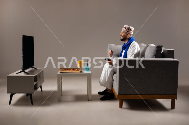تناول وجبات خفيفة اثناء اللعب, قضاء وقت ممتع, بورتريه لرجل عربي خليجي عماني يرتدي الدشداشة والكمه يجلس في غرفة المعيشة على الأريكه ويمسك بيده عصا التحكم, لعب العاب الفيديو بإيماءات تدل على السعادة والسرور، المتعة و التسلية والتفاعل بالبلايستيشن الترفيهية, خلفية رمادية