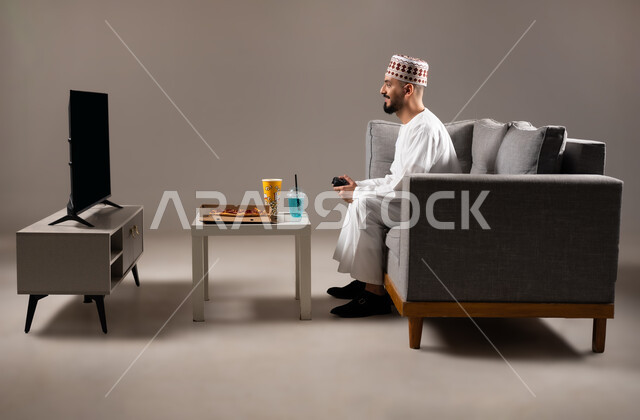 تناول وجبات خفيفة اثناء اللعب, قضاء وقت ممتع, بورتريه لرجل عربي خليجي عماني يرتدي الدشداشة والكمه يجلس في غرفة المعيشة على الأريكه ويمسك بيده عصا التحكم, لعب العاب الفيديو بإيماءات تدل على السعادة والسرور، المتعة و التسلية والتفاعل بالبلايستيشن الترفيهية, خلفية رمادية