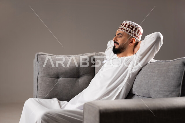 الشعور بالتعب والخمول والحاجة إلى النوم, إيماءات الراحة والإسترخاء، بورتريه لرجل عربي عماني خليجي يرتدي الدشداشة ويضع الكمة  يستريح على الكنبه, تعابير التأمل والتفكير، خلفية رمادية