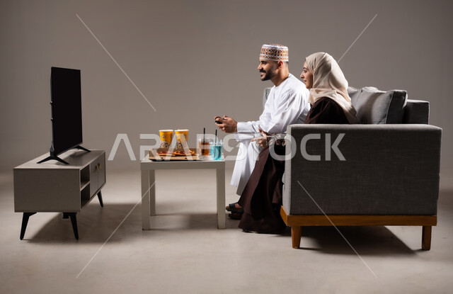 Fun and entertainment with PlayStation entertainment, happy family concept, portrait of an Arab Gulf Omani couple sitting in the living room eating pizza, popcorn and refreshing drinks, having a good time with family and playing video games with gestures indicating happiness and pleasure, gray background