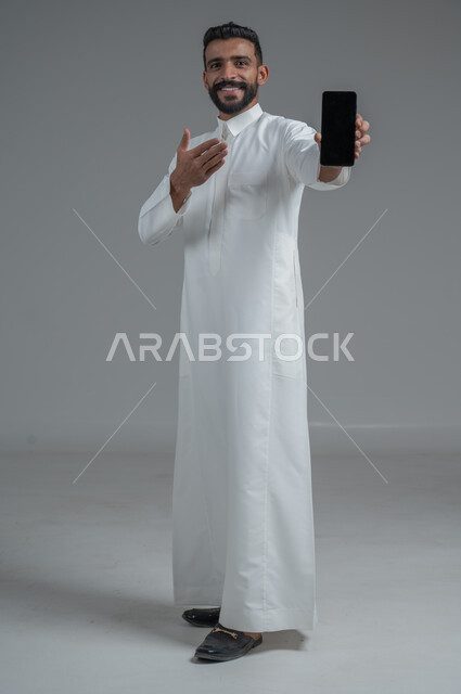 التسوق الالكتروني عبر البرامج والتطبيقات ، بورتريه لرجل عربي خليجي سعودي يرتدي الزي التقليدي يشير بيده الى الهاتف المحمول بشاشة سوداء فارغة ، إيماءات وجه مبتسمة , صورة الجسم بالطول الكامل , خلفية رمادية