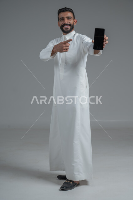 E-shopping through programs and applications, portrait of a Saudi Gulf Arab man wearing traditional clothing, pointing with his hand to a mobile phone with a blank black screen, smiling face gestures, full-length body portrait, gray background