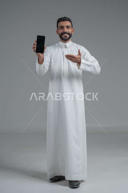 التسوق الالكتروني عبر البرامج والتطبيقات ، بورتريه لرجل عربي خليجي سعودي يرتدي الزي التقليدي يشير بيده الى الهاتف المحمول بشاشة سوداء فارغة ، إيماءات وجه مبتسمة , صورة الجسم بالطول الكامل , خلفية رمادية