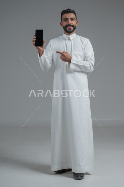 التسوق الالكتروني عبر البرامج والتطبيقات ، بورتريه لرجل عربي خليجي سعودي يرتدي الزي التقليدي يشير بيده الى الهاتف المحمول بشاشة سوداء فارغة ، إيماءات وجه مبتسمة , صورة الجسم بالطول الكامل , خلفية رمادية
