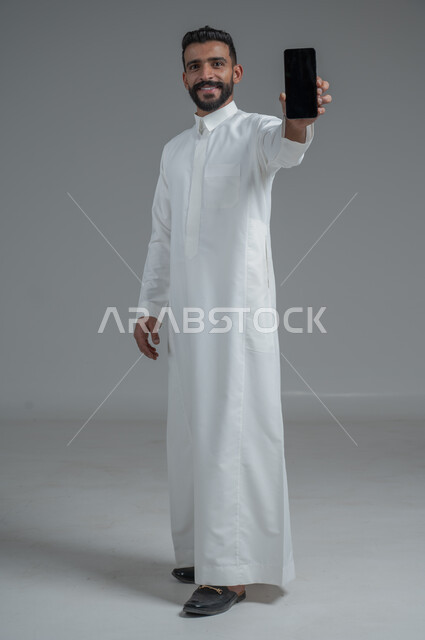 E-shopping through programs and applications, portrait of a Saudi Gulf Arab man wearing traditional clothing and holding a mobile phone in his hand with a blank black screen, smiling face gestures, full-length body portrait, gray background