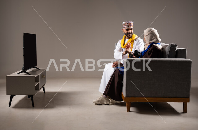 المتعة و التسلية بالبلايستيشن الترفيهية, مفهوم الأسرة السعيدة,  بورتريه لزوجان عربيان خليجيان عمانيان يجلسان في غرفة المعيشة، قضاء وقت ممتع مع العائلة و بلعب ألعاب الفيديو بإيماءات تدل على السعادة والسرور، خلفية رمادية