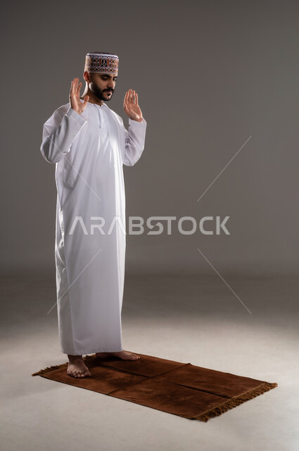 مفهوم أركان الإسلام، العبادة والتقرب إلى الله، إقامة الصلاة على وقتها، أداء الفرائض الدينية الاسلامية، تصوير لرجل عربي خليجي عماني مسلم يرتدي الكمة والدشداشة يصلي, خلفية رمادية