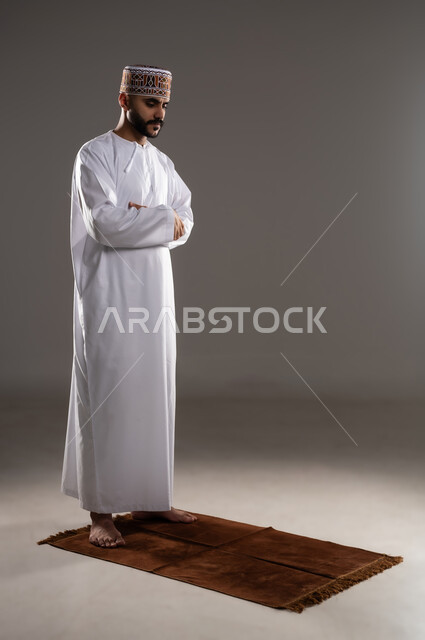 مفهوم أركان الإسلام، العبادة والتقرب إلى الله، إقامة الصلاة على وقتها، أداء الفرائض الدينية الاسلامية، بورتريه لرجل عربي خليجي عماني مسلم يرتدي الكمة والدشداشة يصلي, صورة الجسم بالطول الكامل, خلفية رمادية