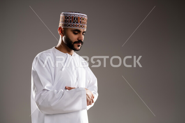 الإجتهاد في العباده في شهر رمضان المبارك, مفهوم أركان الإسلام والتقرب إلى الله، إقامة الصلاة على وقتها، أداء الفرائض الدينية الاسلامية، بورتريه لرجل عربي خليجي عماني مسلم يرتدي الكمة والدشداشة ويصلي,  خلفية رمادية
