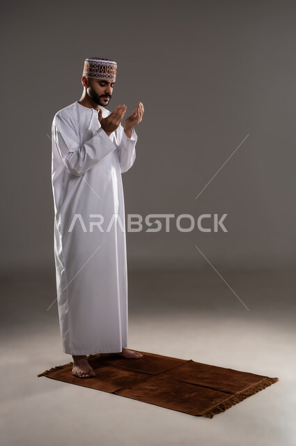 مفهوم التضرع والتعبد، الإجتهاد في العباده في شهر رمضان المبارك, بورتريه لرجل عربي خليجي عماني مسلم يرتدي الكمة والدشداشة يقف على سجادة الصلاة ويرفع يديه ويدعي, الإكثار من ذكر الله تعالى من خلال الإستغفار والدعاء, صورة الجسم بالطول الكامل, خلفية رمادية