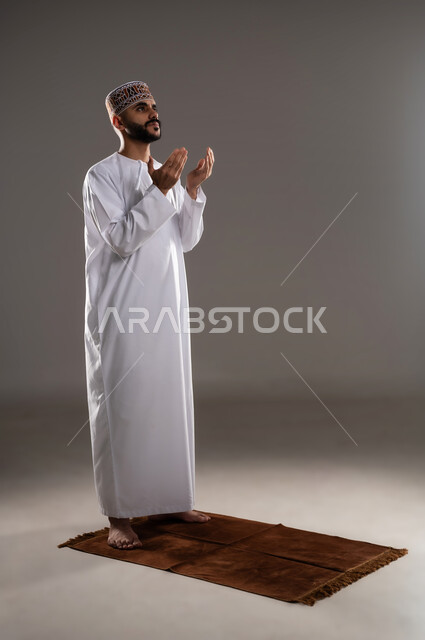 مفهوم التضرع والتعبد، الإجتهاد في العباده في شهر رمضان المبارك, بورتريه لرجل عربي خليجي عماني مسلم يرتدي الكمة والدشداشة يقف على سجادة الصلاة ويرفع يديه ويدعي, الإكثار من ذكر الله تعالى من خلال الإستغفار والدعاء, صورة الجسم بالطول الكامل, خلفية رمادية