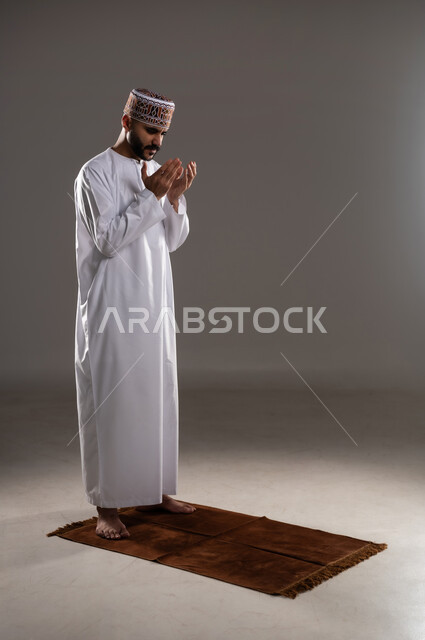 مفهوم التضرع والتعبد، الإجتهاد في العباده في شهر رمضان المبارك, بورتريه لرجل عربي خليجي عماني مسلم يرتدي الكمة والدشداشة يقف على سجادة الصلاة ويرفع يديه ويدعي, الإكثار من ذكر الله تعالى من خلال الإستغفار والدعاء, صورة الجسم بالطول الكامل, خلفية رمادية