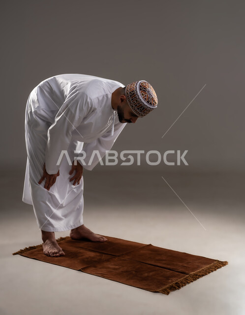 Diligence in worship during the blessed month of Ramadan, the concept of the pillars of Islam and getting closer to God, performing prayers on time, performing Islamic religious duties, a portrait of an Arab Gulf Omani Muslim man wearing a kuma and dishdasha and kneeling on the prayer rug, gray background