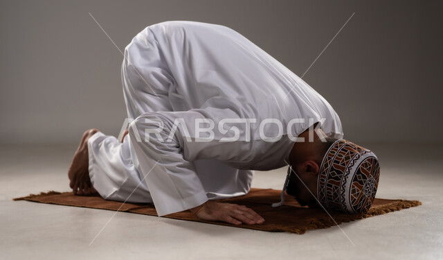 الإجتهاد في العباده في شهر رمضان المبارك, مفهوم أركان الإسلام والتقرب إلى الله، إقامة الصلاوات على وقتها، أداء الفرائض الدينية الاسلامية، بورتريه لرجل عربي خليجي عماني مسلم يرتدي الكمة والدشداشة ويسجد على سجادة الصلاة,  خلفية رمادية