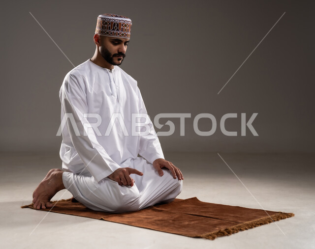 الإجتهاد في العباده في شهر رمضان المبارك, مفهوم أركان الإسلام والتقرب إلى الله، إقامة الصلاة على وقتها، أداء الفرائض الدينية الاسلامية، بورتريه لرجل عربي خليجي عماني مسلم يرتدي الكمة والدشداشة ويصلي,  خلفية رمادية