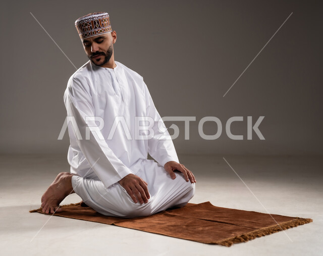 الإجتهاد في العباده في شهر رمضان المبارك, مفهوم أركان الإسلام والتقرب إلى الله، إقامة الصلاة على وقتها، أداء الفرائض الدينية الاسلامية، بورتريه لرجل عربي خليجي عماني مسلم يرتدي الكمة والدشداشة ويصلي,  خلفية رمادية