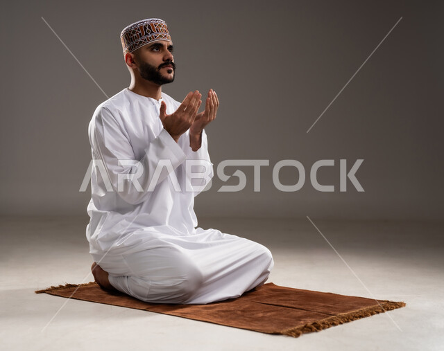 مفهوم التضرع والتعبد، الإجتهاد في العباده في شهر رمضان المبارك, بورتريه لرجل عربي خليجي عماني مسلم يرتدي الكمة والدشداشة يجلس على سجادة الصلاة ويرفع يديه ويدعي, الإكثار من ذكر الله تعالى من خلال الإستغفار والدعاء, خلفية رمادية