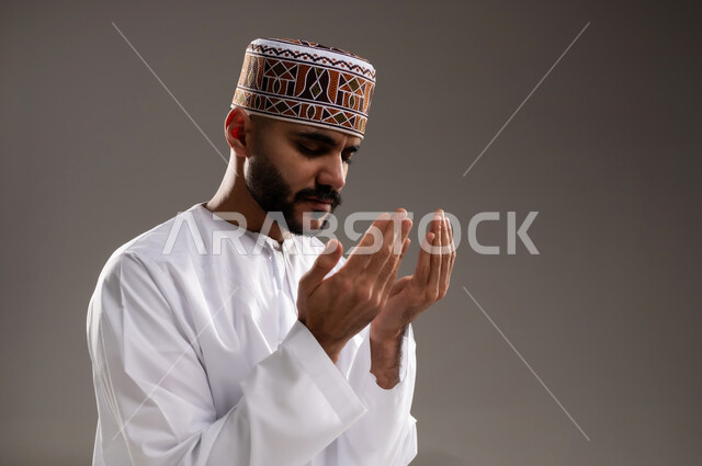 The concept of supplication and worship, diligence in worship in the holy month of Ramadan, close-up portrait of an Arab Gulf Omani Muslim man wearing a kabbah and dishdasha, sitting on the prayer rug, raising his hands and praying, increasing the remembrance of God Almighty through seeking forgiveness and supplication, gray background