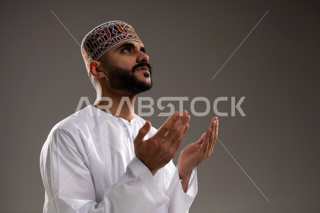 مفهوم التضرع والتعبد، الإجتهاد في العباده في شهر رمضان المبارك, بورتريه مقرب لرجل عربي خليجي عماني مسلم يرتدي الكمة والدشداشة يجلس على سجادة الصلاة ويرفع يديه ويدعي, الإكثار من ذكر الله تعالى من خلال الإستغفار والدعاء, خلفية رمادية