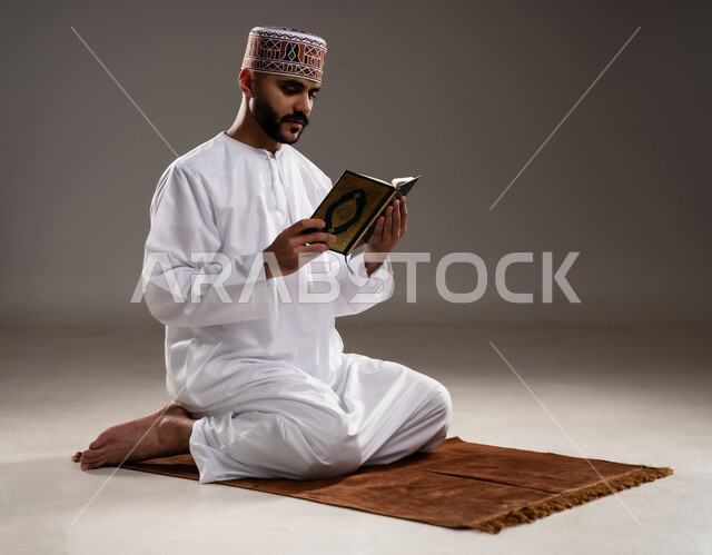 مفهوم قراءة القران، الإجتهاد في العباده والتقرب الى الله في شهر رمضان المبارك, بورتريه لرجل عربي خليجي عماني مسلم يرتدي الكمة والدشداشة يجلس على سجادة الصلاة ويقرأ المصحف الشريف, خلفية رمادية